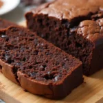 Sliced brownie bread loaf surrounded by chocolate chips and cocoa powder.