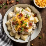 Close-up of creamy baked potato salad with cheddar, bacon, and green onions in a white bowl on rustic wood background.