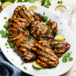 Juicy grilled chicken thighs on a white serving platter with fresh cilantro, lime wedges, and charred grill marks.