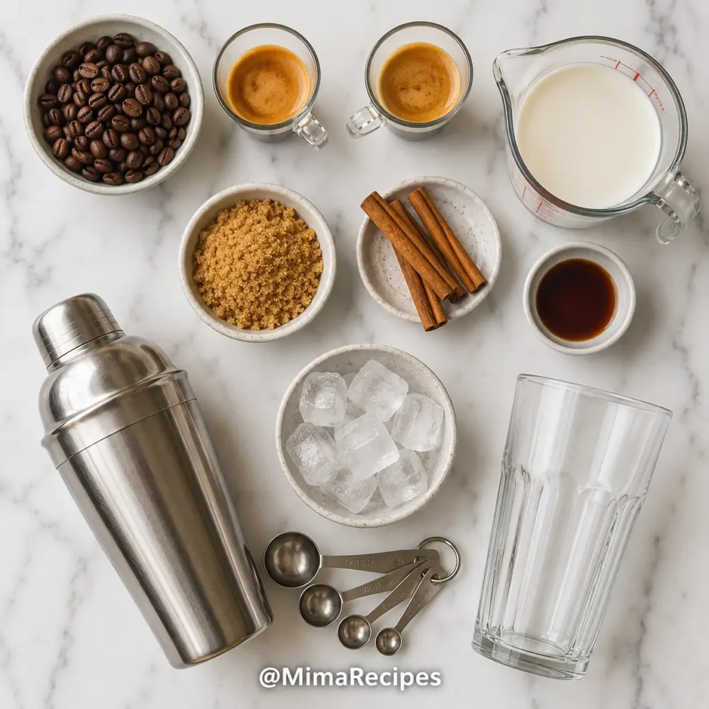 Flat lay of espresso beans, espresso shots, brown sugar, cinnamon sticks, vanilla extract, oat milk, ice cubes, cocktail shaker, measuring spoons, and a tall glass arranged on a white marble countertop.